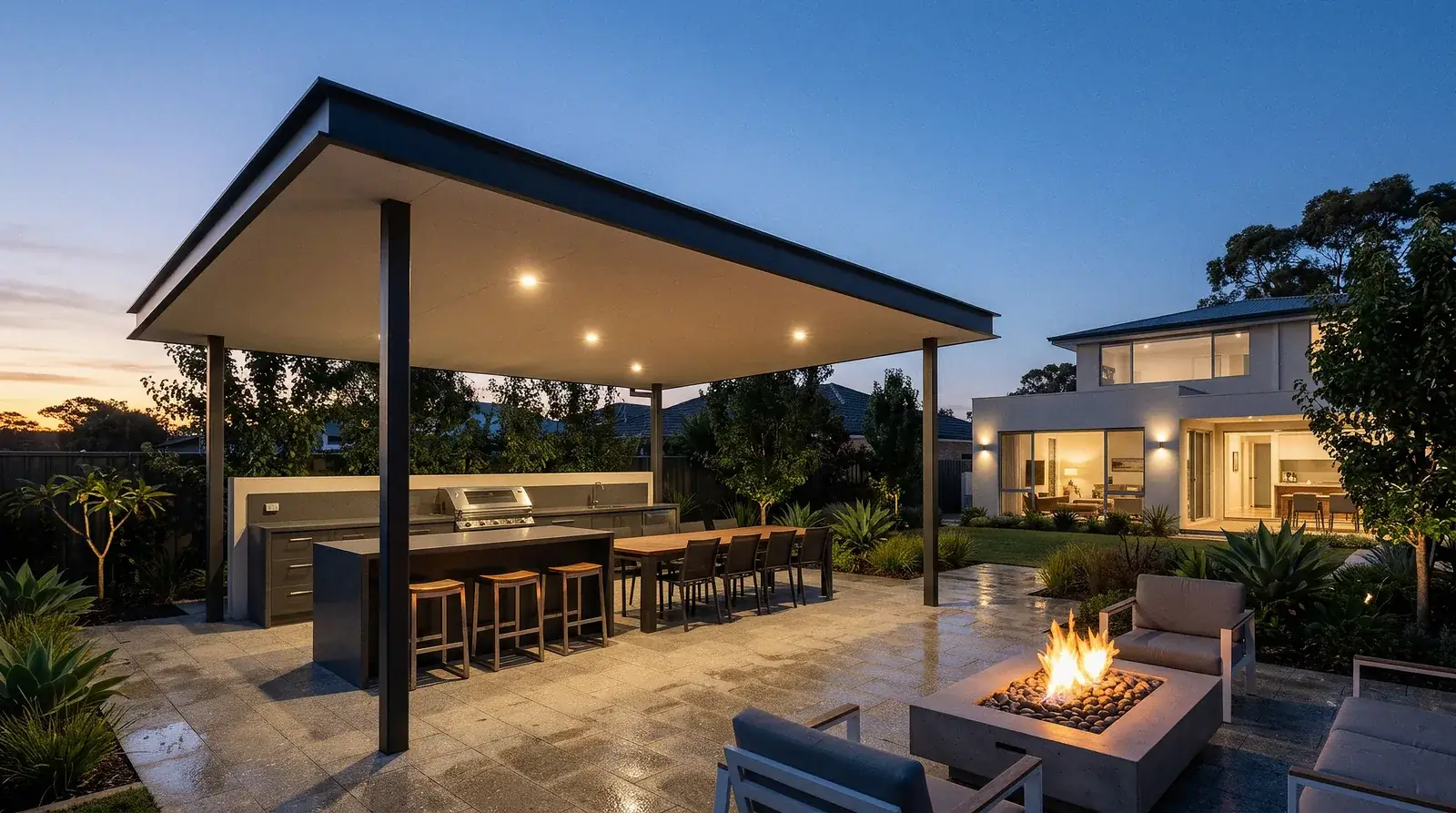 A freestanding flat roof patio lit at dusk over a built-in outdoor kitchen and a long dining table, downlights set flush into its ceiling and the house glowing separately in the distance